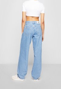 Person som bär ljusblå vida jeans och en vit kortärmad t-shirt, stående mot en enfärgad ljusgrå bakgrund.