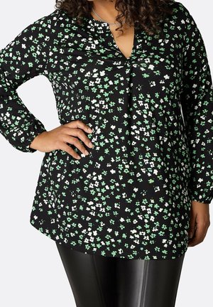 Femme portant un haut noir à manches longues avec un petit motif floral vert et blanc et des leggings noirs aspect cuir, main sur la hanche.