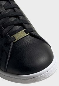 Schwarze Leder-Sneaker mit rundem Zehenbereich, strukturiertem Obermaterial, schwarzen Schnürsenkeln und einem metallisch goldenen Akzent im Schnürsystem. Weiße Gummisohle.