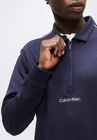 Άτομο που φοράει μπλε σκουρόχρωμο φούτερ Calvin Klein με φερμουάρ μέχρι το λαιμό, χέρι που ρυθμίζει το φερμουάρ κοντά στον γιακά, μερικό πρόσωπο ορατό, λευκό πουκάμισο από κάτω.