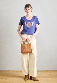 Μωβ μπλούζα με πολύχρωμο γραφικό φτερού, κρεμ παντελόνι με φαρδιές γάμπες και καφέ τσάντα με επένδυση και κοντές λαβές. Καφέ μποτάκια με τακούνι.