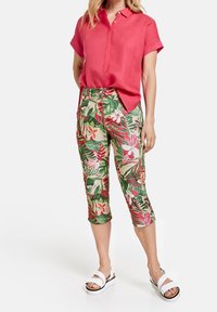 Punainen lyhythihainen paita yhdistettynä vihreisiin caprihousuihin, joissa on vaaleanpunaisia ja valkoisia kukkia. Valkeat sandaalit viimeistelevät asun.