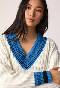 Weißer gerippter Pullover mit tiefem V-Ausschnitt und blauen sowie schwarzen Akzenten, gestreiften Details an den Bündchen. Weicher Stoff, lässige Passform.