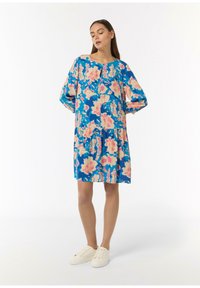 Blumenkleid in Blau mit rosa und pfirsichfarbenen Mustern, runder Ausschnitt, lockere Ärmel und ein gestuftem Rock. Kombiniert mit weißen Sneakers.