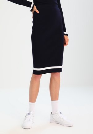 Femme portant une jupe bleu marine jusqu'au genou avec bordure blanche, chaussettes blanches et baskets blanches Reebok, debout avec une main sur la hanche.