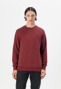 Maroon sweatshirt with a round neck, long sleeves, ribbed cuffs, and a small black logo on the chest. Smooth texture and fitted design.