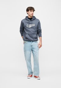 Νεαρός άνδρας που φοράει μπλε φούτερ Converse, ανοιχτόχρωμα μπλε τζιν και κόκκινα αθλητικά παπούτσια, στέκεται μπροστά σε ένα απλό λευκό φόντο.