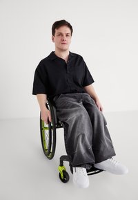 Svart kortermet skjorte, grå løse jeans og hvite joggesko, båret av en person i rullestol med grønne detaljer på hjulene.