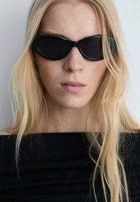 Ochelari de soare negri în formă de ochi de pisică, cu un finisaj lucios și rame subțiri. Modelul are păr lung blond și poartă un top negru cu umerii descoperiți.