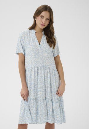 Žena s dlhými hnedými vlasmi, oblečená v svetlomodrých a bielych krátkych rukávoch so vzorovanými vrstvenými šatami na jednofarebnom pozadí.