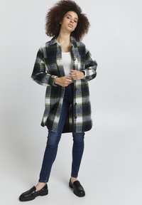 Plaid oversized shirt jacket in dark green, navy, and white; button closure, long sleeves, and two front pockets. Worn over a white top.
