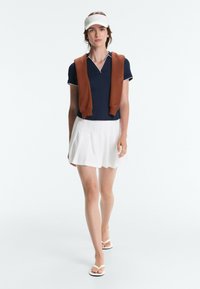 Femme marchant portant un polo bleu marine, une jupe blanche, une visière blanche, un pull marron drapé sur les épaules et des sandales blanches type tongs.