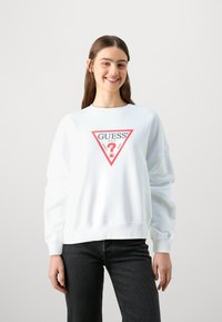 Biela mikina s dlhými rukávmi a rebrovanými manžetami a spodným lemom, na prednej strane s trojuholníkovým červeným a čiernym logom "GUESS".