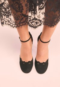 Sorte ruskindsstiletter med bølgede kanter og udskårne detaljer, med en tynd ankelrem. Sat mod en lys pink baggrund.