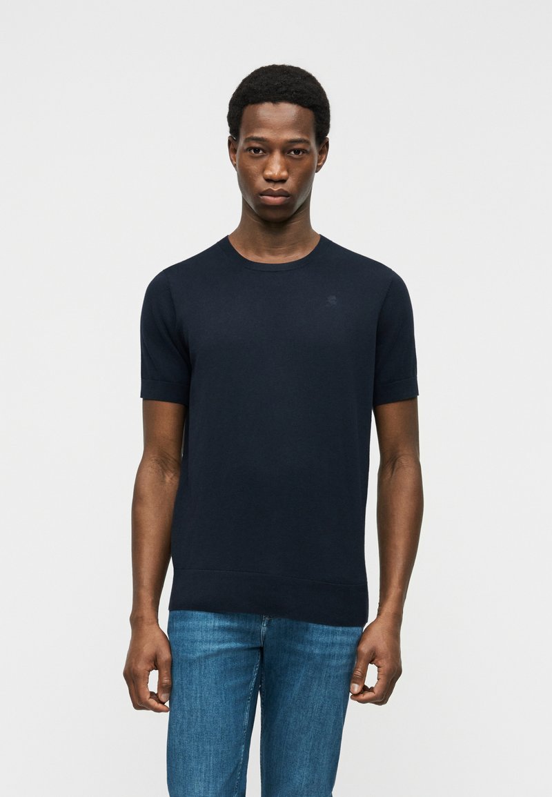 Jeune homme aux cheveux courts portant une chemise bleu marine à manches courtes et un jean bleu, debout devant un fond blanc uni.