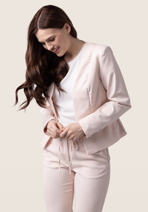 Světle růžový blazer a odpovídající kalhoty vyrobené z hladké tkaniny, které mají přizpůsobený střih a jemnou gumu v pase.