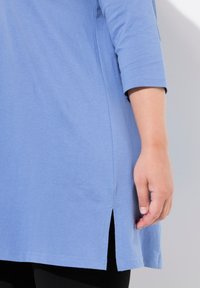 Helles Blau, Tunika-Oberteil mit kurzen Ärmeln und abgerundetem Saum. Verfügt über seitliche Schlitze am unteren Ende für Bewegungsfreiheit und eine glatte Baumwollstruktur.