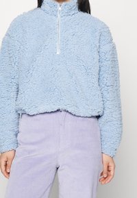 Ljusblå fleece-pullover med hög krage och halvdragkedja. Texturerad, fluffig yta; bärs med lavendelfärgade kordbyxor.