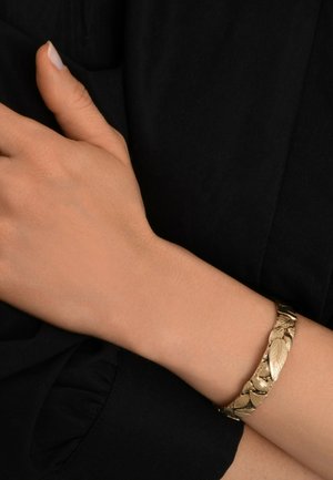 Bracelet avec un motif en feuille d'or sur un poignet, présentant un design texturé avec une finition lisse. La main repose sur un fond en tissu noir.