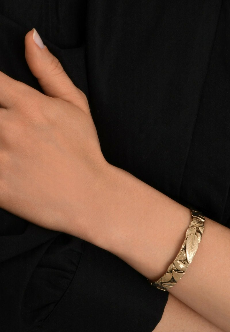 Bracelet avec un motif en feuille d'or sur un poignet, présentant un design texturé avec une finition lisse. La main repose sur un fond en tissu noir.
