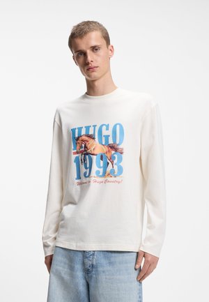Jeune homme portant une chemise blanche à manches longues avec un graphisme de cheval et le texte "HUGO 1993", associé à un jean bleu clair.