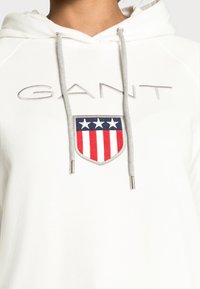 Sudadera blanca con cordones grises y un emblema en forma de escudo en rojo, blanco y azul con tres estrellas blancas sobre rayas horizontales.