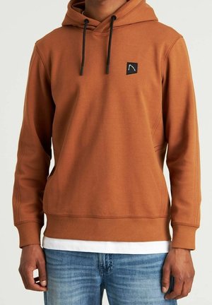 Mann trägt rostfarbenen Kapuzenpullover mit schwarzen Kordeln, kleinem schwarzem Logopatch auf der Brust, weißem Unterhemd und blauer Jeans.