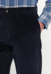 Marineblauwe ribfluwelen broek met zichtbare verticale ribbels, voorzien van een voorzak en een knoopsluiting. Gecombineerd met een blauw geruit overhemd.