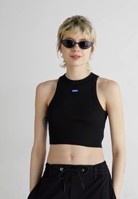 Black cropped tank top with a rounded neckline and a blue logo. The model wears black sunglasses and high-waisted black pants.