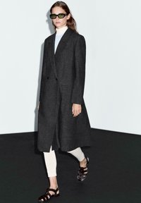 Cappotto lungo grigio con un design strutturato, davanti doppiopetto, abbinato a leggings bianchi aderenti e sandali con cinturini marroni. Occhiali da sole scuri indossati.