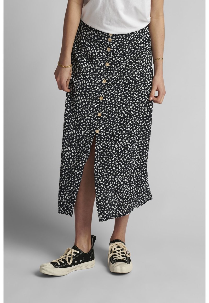 Femme portant une jupe midi noire à fleurs avec des boutons sur le devant et une fente à l'avant, un T-shirt blanc, des baskets basses noires en toile et des bracelets dorés.