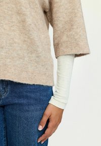 Bež pehmea kudumiga sviiter lühikeste varrukatega, mille all on pikkade varrukatega valge topp. Seljas tume sinine denimist teksapaar.