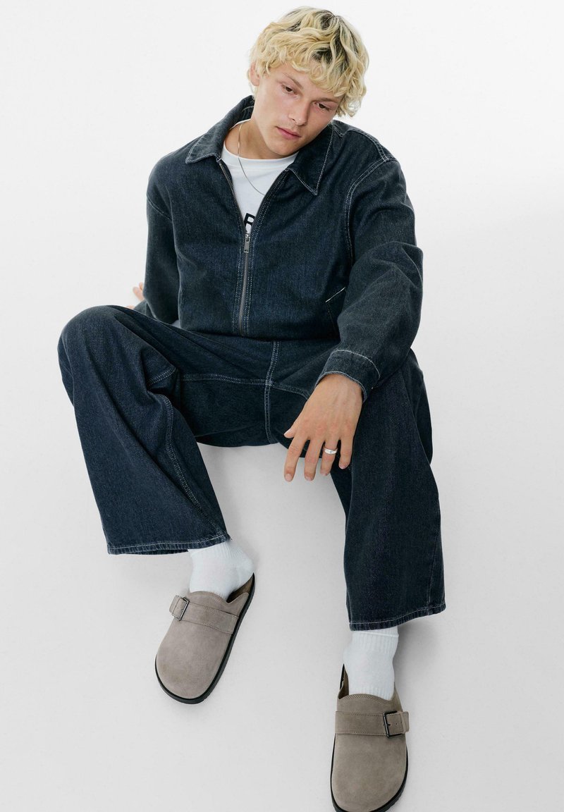Tuta in denim blu scuro con cuciture bianche, chiusura a zip frontale e vestibilità rilassata. Indossa zoccoli in suede grigi con fibbia e calzini bianchi.