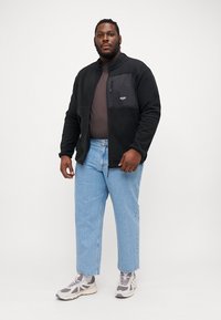 Black fleece jacket with a zip pocket, worn over a brown top, paired with light blue jeans and gray sneakers. Model poses frontally.