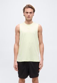 Jeune homme debout, portant un débardeur jaune clair Puma et un short noir Puma, sur un fond clair uni.