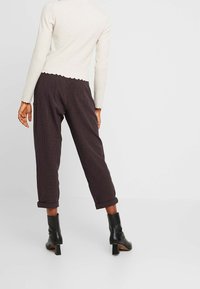 Cremfarbener, gerippter Langarmoberteil mit gewelltem Saum, kombiniert mit braunen, karierten Hosen, hochgekrempelten Ärmeln und schwarzen, hochhackigen Ankle Boots.