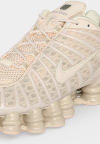 Κοντινή λήψη σε αθλητικό παπούτσι Nike Shox μπεζ χρώματος με υφασμάτινο επάνω μέρος με υφή, ριμπ ύφασμα και διακριτές αποσβεστικές κολόνες στη σόλα.