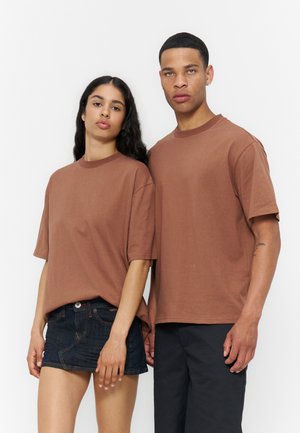 Két álló felnőtt, akik egyező barna oversized pólót viselnek; az egyik sötét farmer szoknyával, a másik fekete nadrággal, egyszínű háttér előtt.