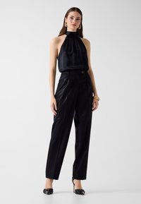 Frau trägt ein schwarzes Neckholder-Top, hoch taillierte schwarze Hose, schwarze High Heels, goldene Creolen und ein goldenes Armband, steht vor weißem Hintergrund.