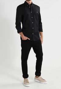 Camicia nera a maniche lunghe con bottoni e colletto, abbinata a pantaloni neri arrotolati e sneakers beige chiaro, presentata da un modello in piedi.