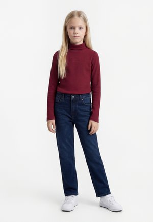 Fille blonde debout, face à nous, portant un col roulé bordeaux, un jean bleu foncé et des baskets blanches sur un fond blanc uni.