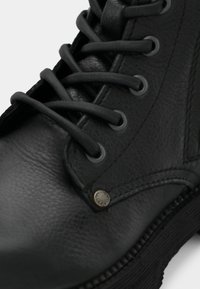 Close-up van een zwart leren laars met dikke veters, metalen oogjes, stiksels en een rubberen zool met textuur.