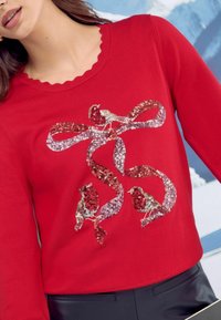 Punainen neulepaita, jossa on viistetty pääntie, jossa on strassikuvio kahdesta linnusta ja nauhoista punaisen ja hopean sävyissä. Silkkinen pinta.