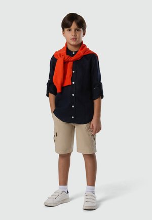 Junge steht und trägt ein navyblaues Hemd zum Knöpfen, beige Cargo-Shorts, weiße Turnschuhe und einen orangen Pullover, der über seine Schultern gelegt ist.