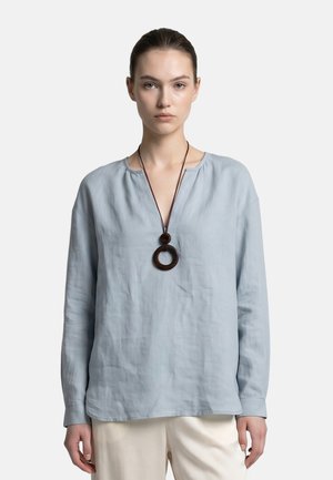 Jonge vrouw met bruin haar in een knot, die een lichtblauw blouse met lange mouwen, crèmekleurige broek en een ketting met een grote ronde hanger draagt.