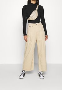 Beige weit geschnittene Hose mit Falten, kombiniert mit einem schwarzen Langarmoberteil und einer hellbraunen Umhängetasche mit mehreren Reißverschlusstaschen.
