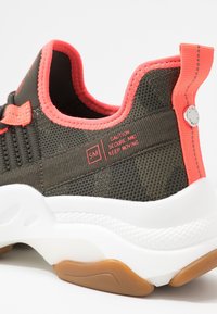 Tummanvihreä verkkokenkä koralliakseilla, pehmustettu valkoinen pohja ja heijastava yksityiskohta. Varustettu tarralla, jossa lukee "Varoitus: Varmista ja jatka liikettä."