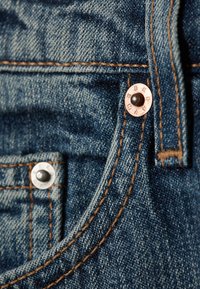 Sinine teksapükste tasku suumiga, kus on nähtavad vase- ja hõbesepad ning oranži niidiga õmblused.