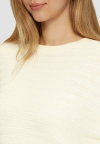 Pull en tricot couleur crème avec une texture côtelée, un col rond et des fibres douces. Des lignes horizontales visibles ajoutent un détail subtil.