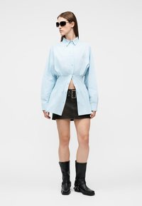 Femme portant une chemise bleu clair cintrée à la taille, une jupe ceinturée noire, des bottes noires mi-mollet et des lunettes de soleil foncées, debout devant un fond blanc.